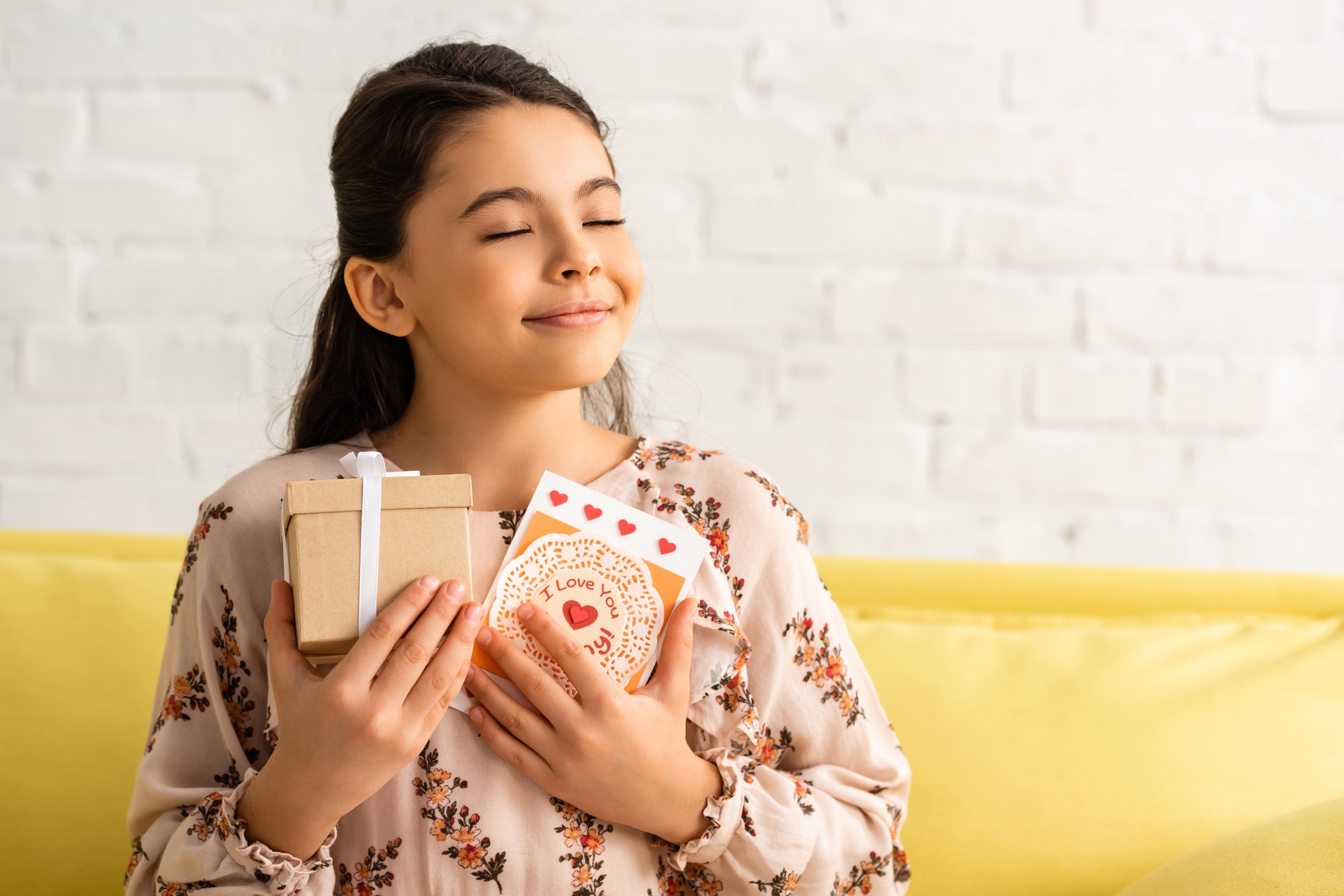 happy child in elegant dress sitting with closed eyes and holding gift box and mothers day card with