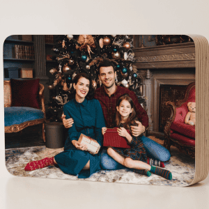 Taco ovalado personalizado de Navidad con fotografía familiar impresa, acabado mate y grosor de 19 mm, colocado sobre una mesa navideña con decoración de luces cálidas, ramas de pino, bolas rojas y lazos, destacando su diseño moderno y elegante como regalo y recuerdo navideño.